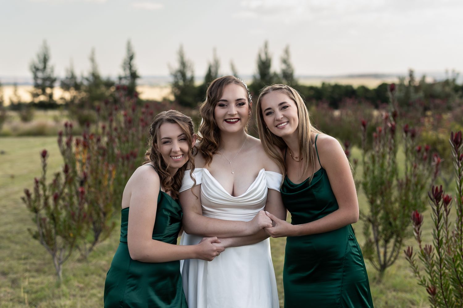 Wedding party in emerald and white dresses embrace while posing in garden setting.