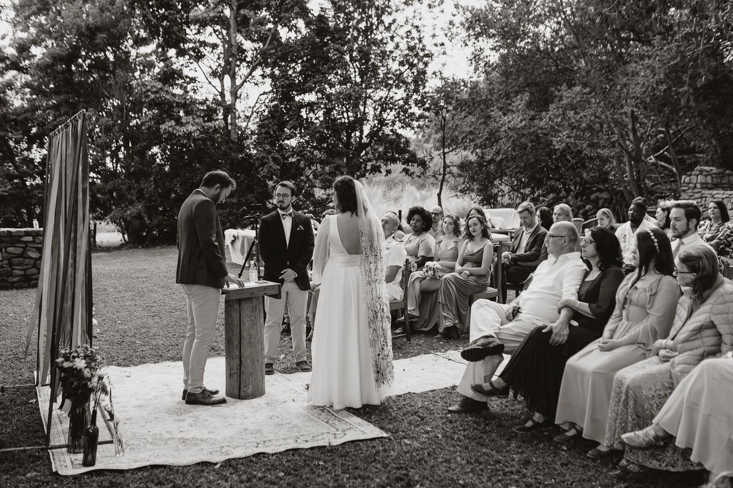 A black and white photo captures a wedding ceremony in a garden setting with seated guests.