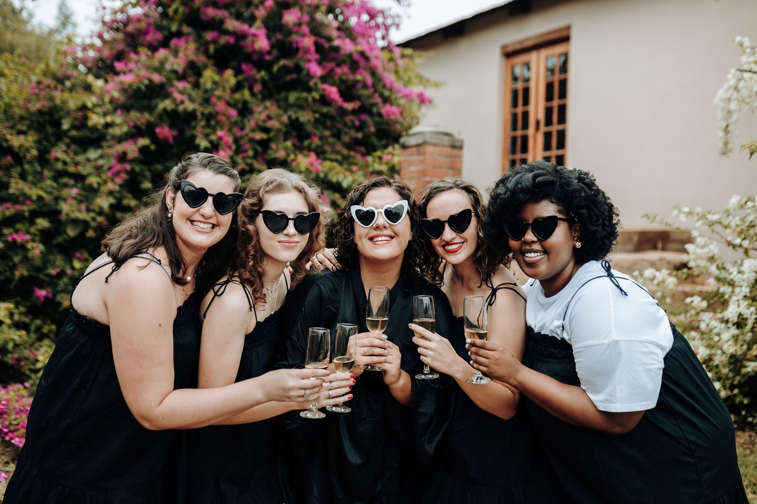Wedding party in black dresses and sunglasses toasting with champagne in garden.