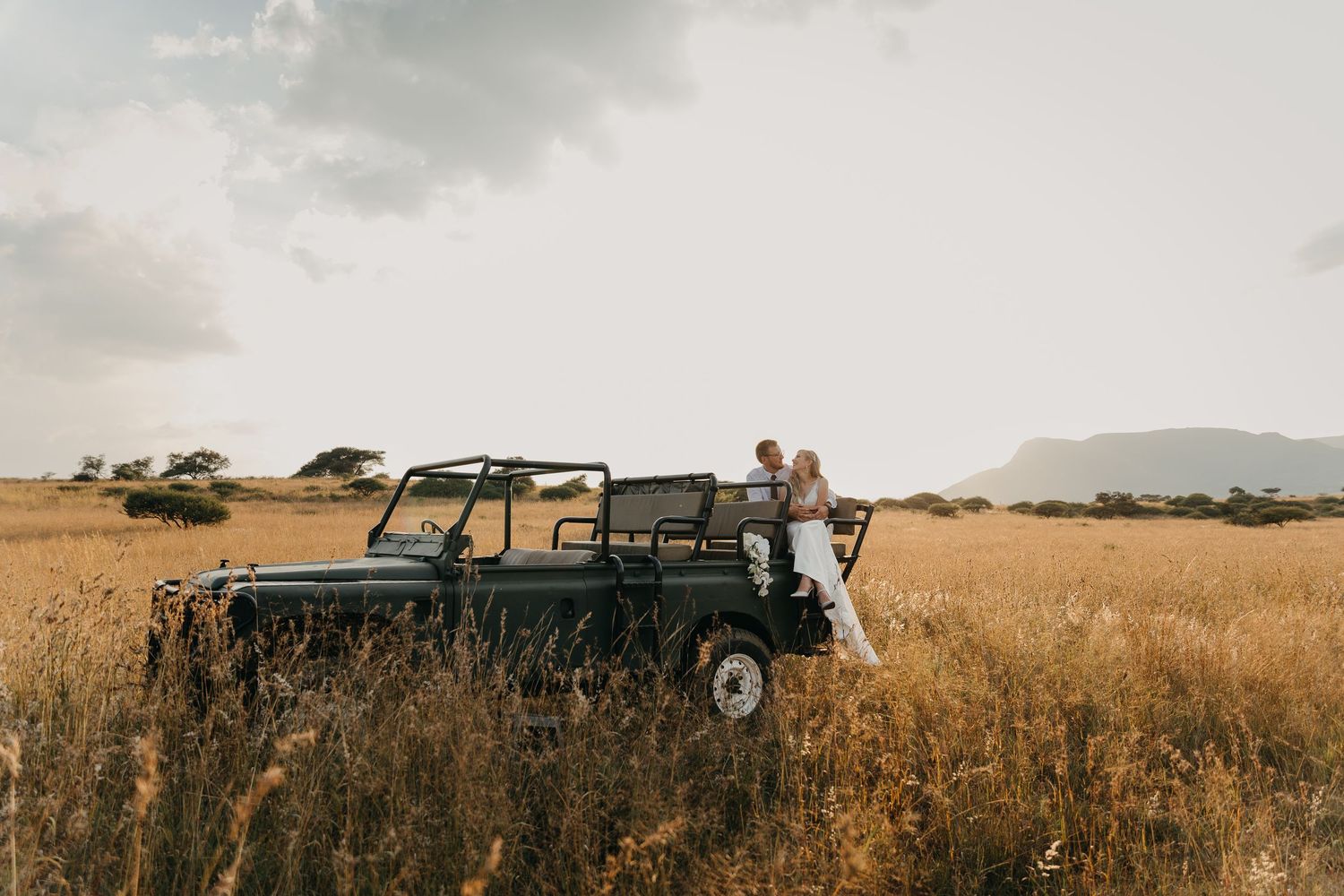 Newlyweds pose with vintage Land Rover in golden African savanna landscape.