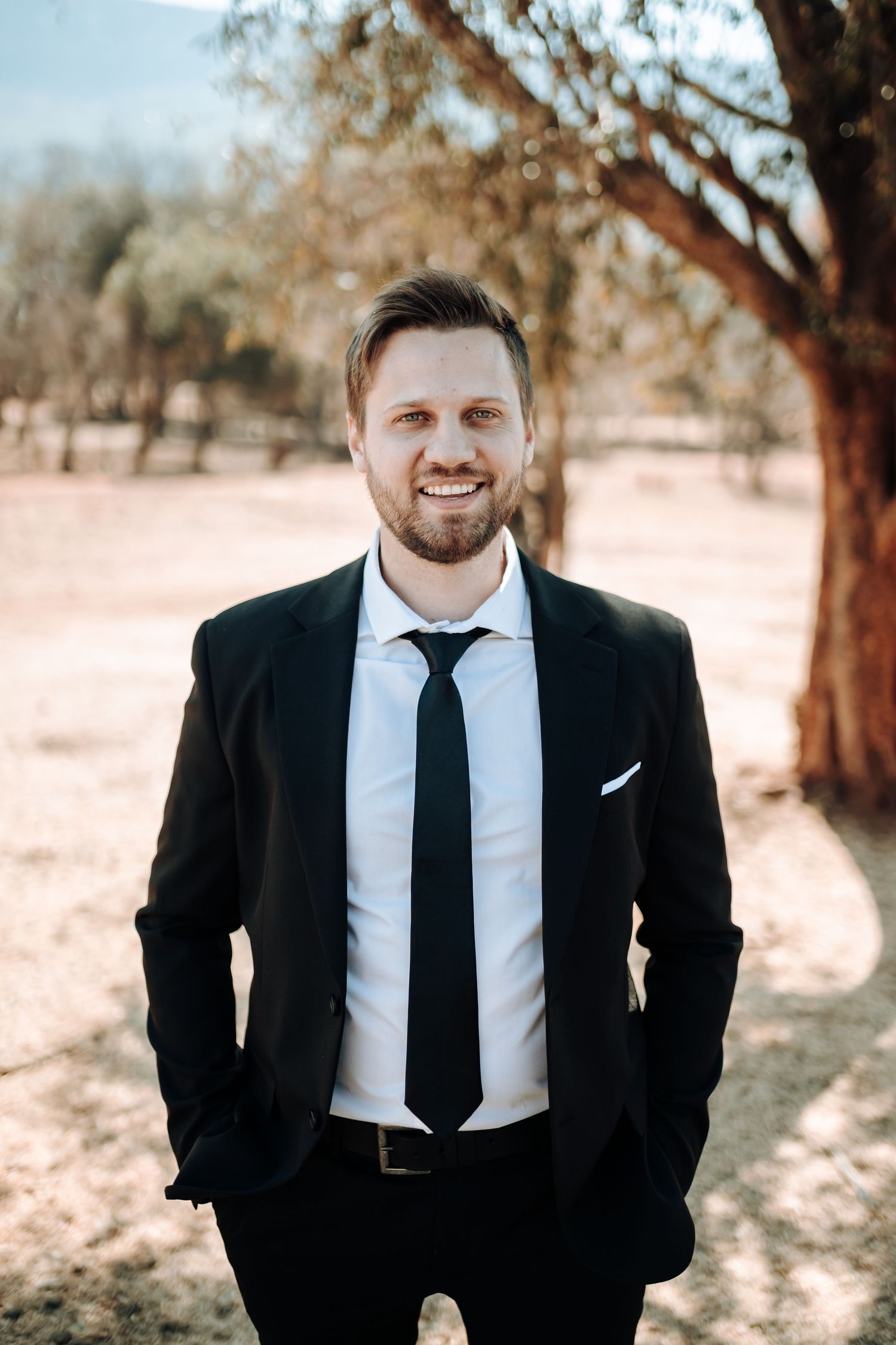 Wedding party member in dark suit poses outdoors against desert background.