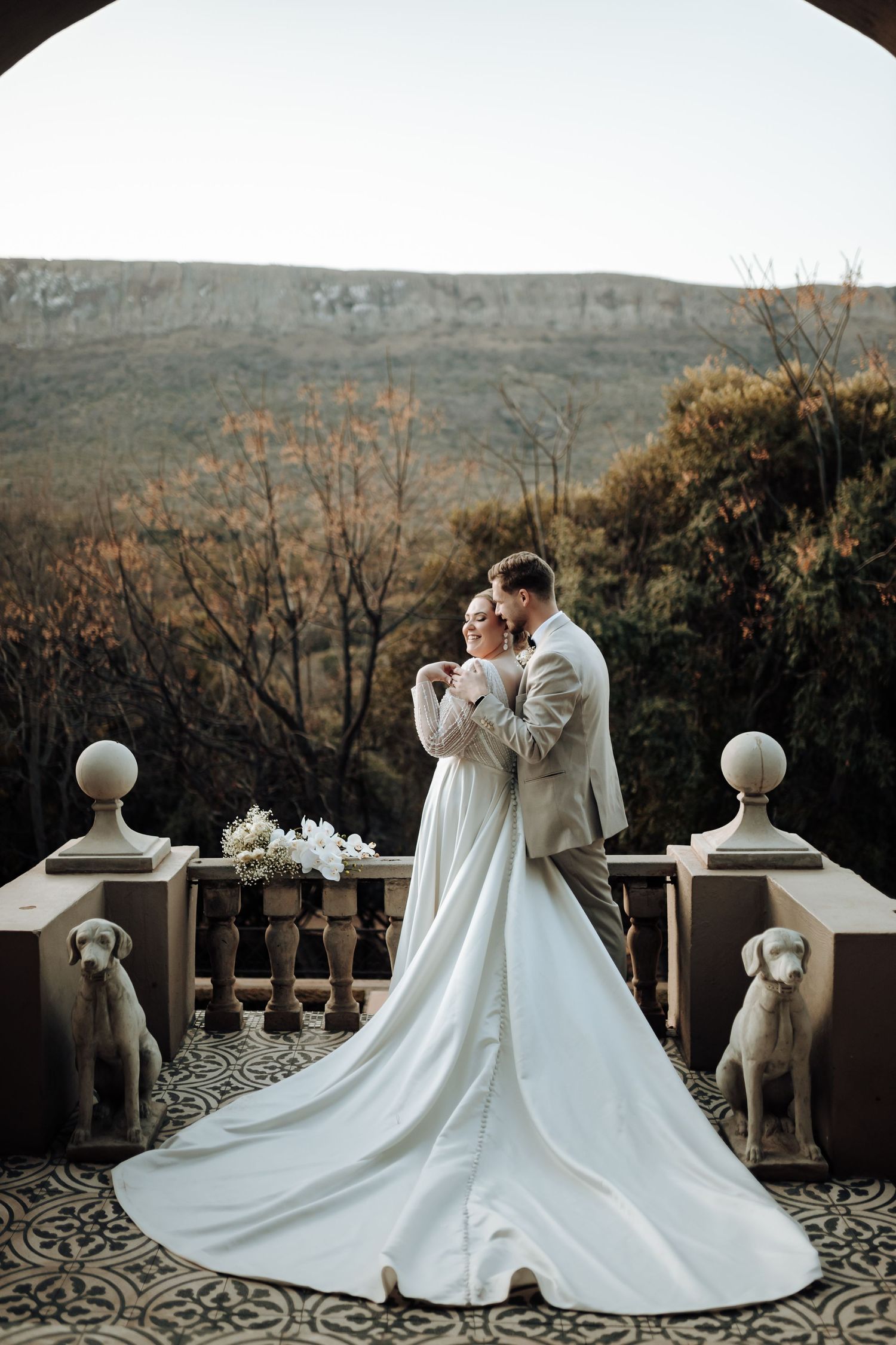 Romantic wedding portrait on stone terrace with decorative statues and mountain background.