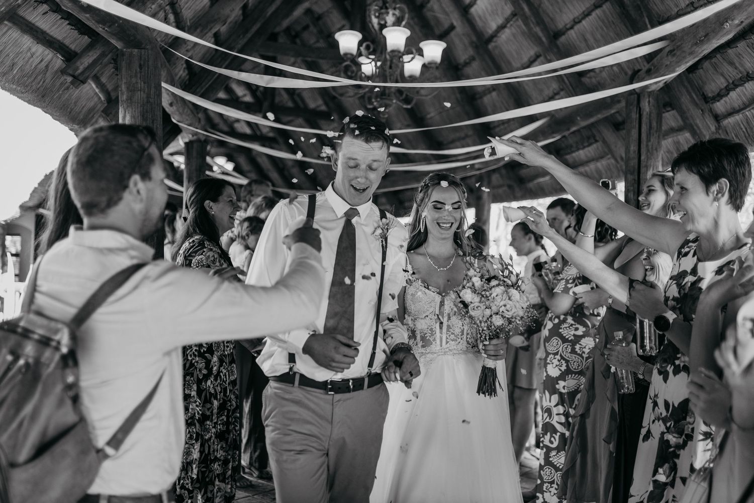 Black and white photo of wedding celebration under rustic wooden beams with guests cheering.
