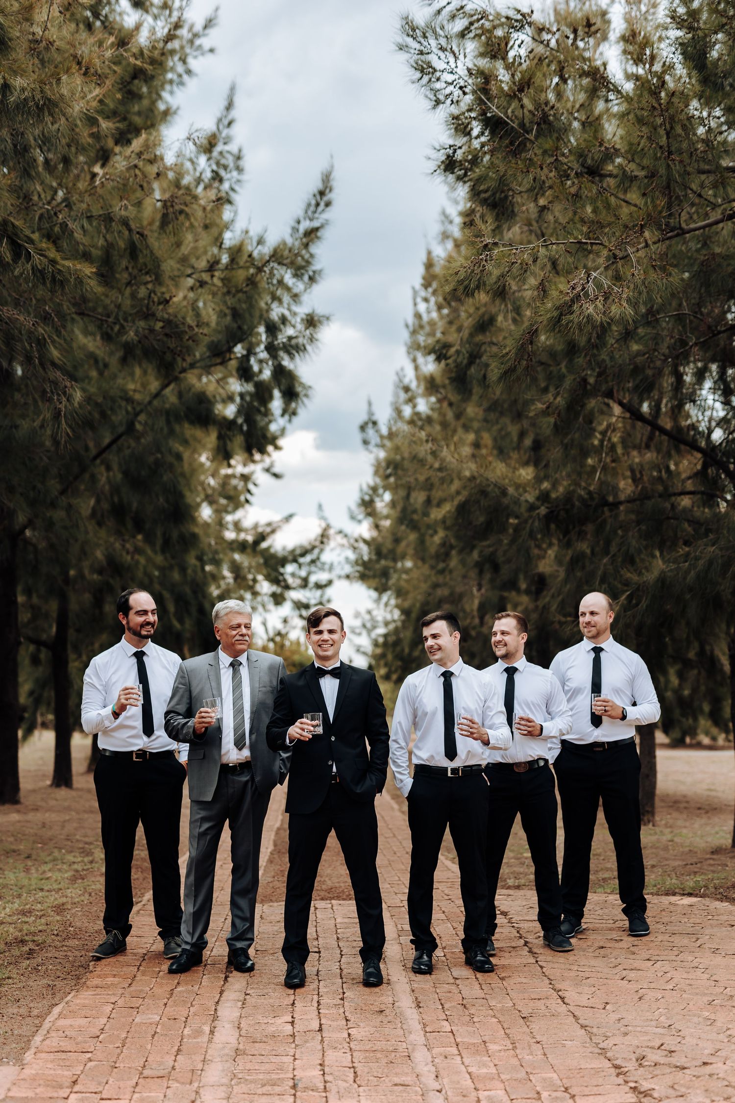 Wedding party members stand in formation on a brick path surrounded by tall pine trees.
