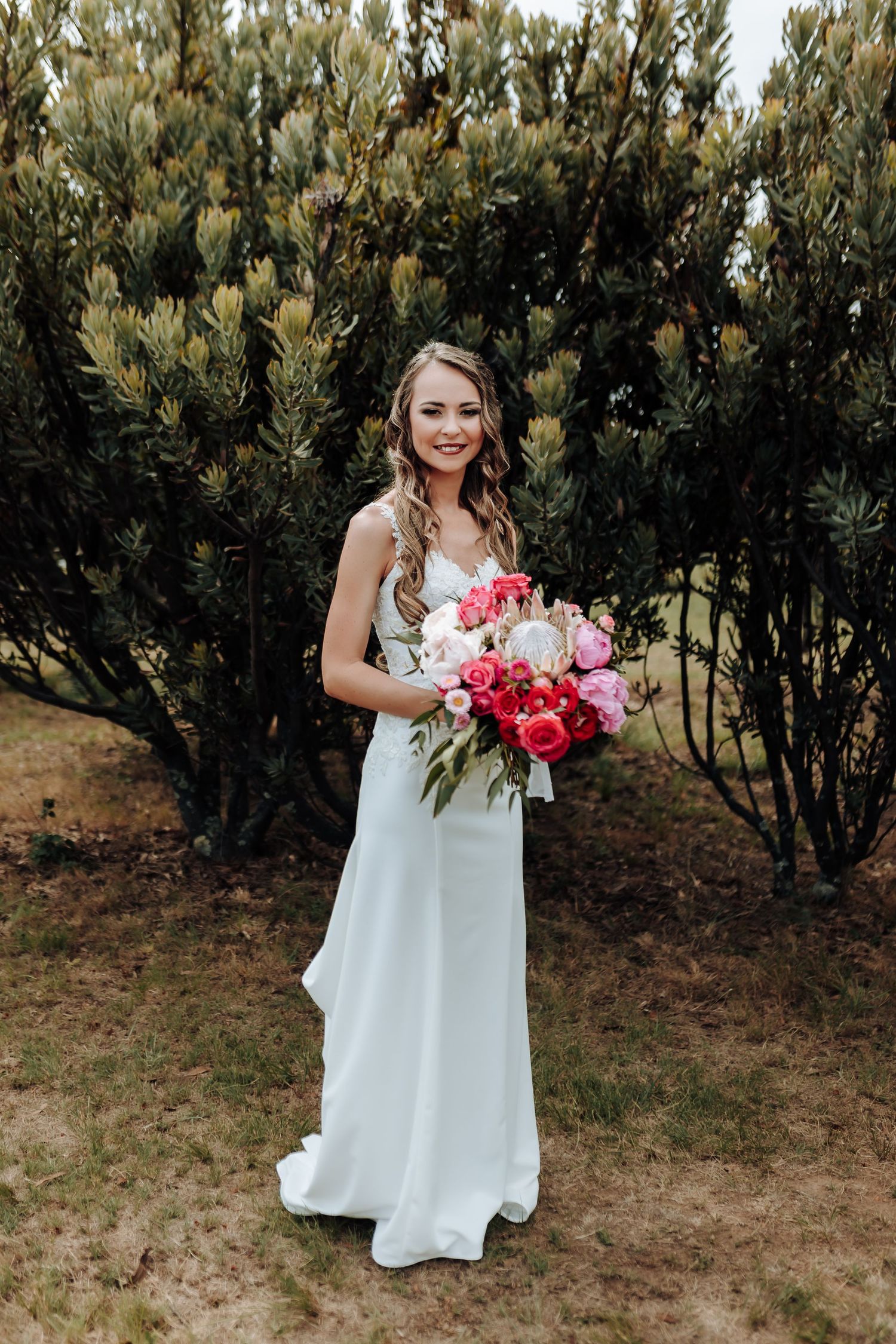 Bride in elegant white gown holds vibrant pink and red bouquet in front of garden shrubs.