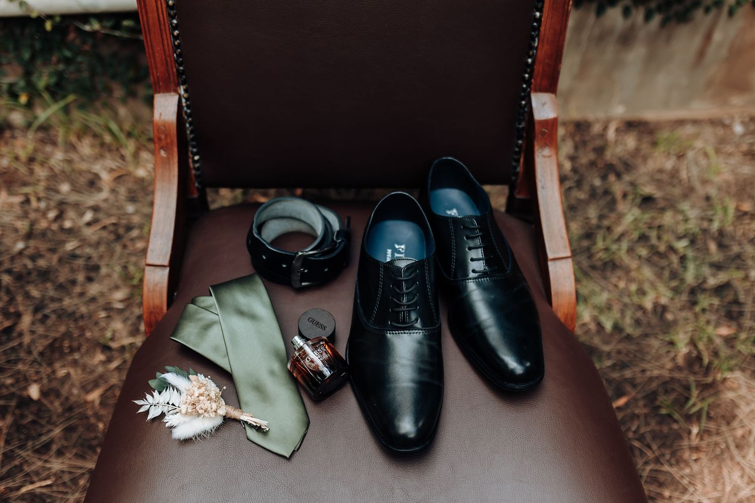 Wedding accessories including black dress shoes, sage green tie and boutonniere arranged on brown leather chair.