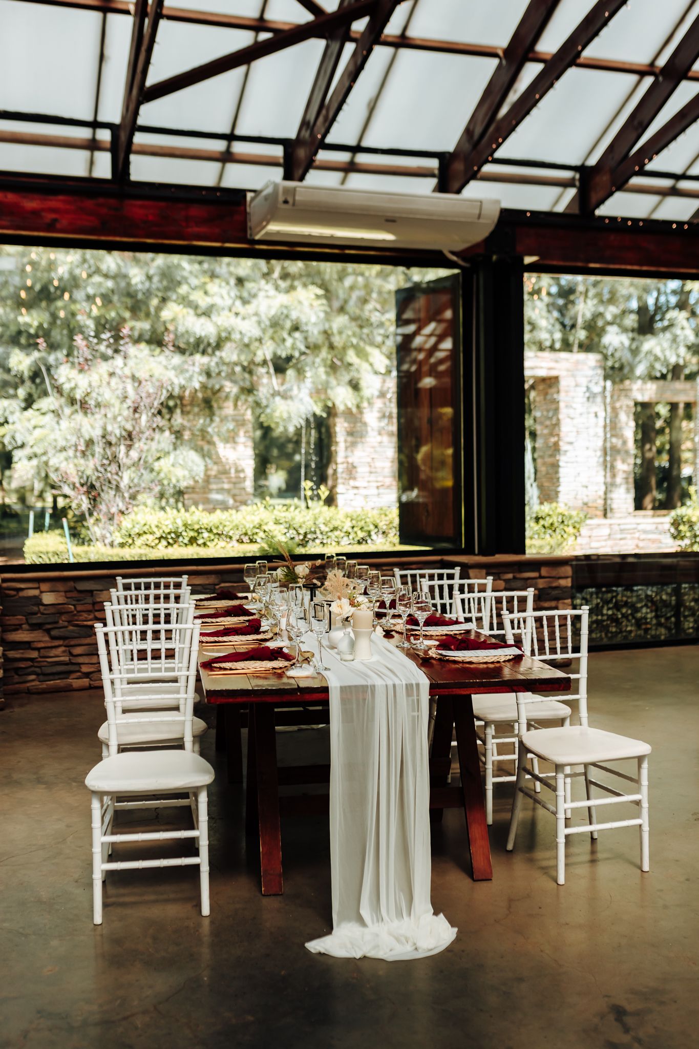 Wedding dining setup with white chairs and flowing fabric runner under a glass-paneled roof.