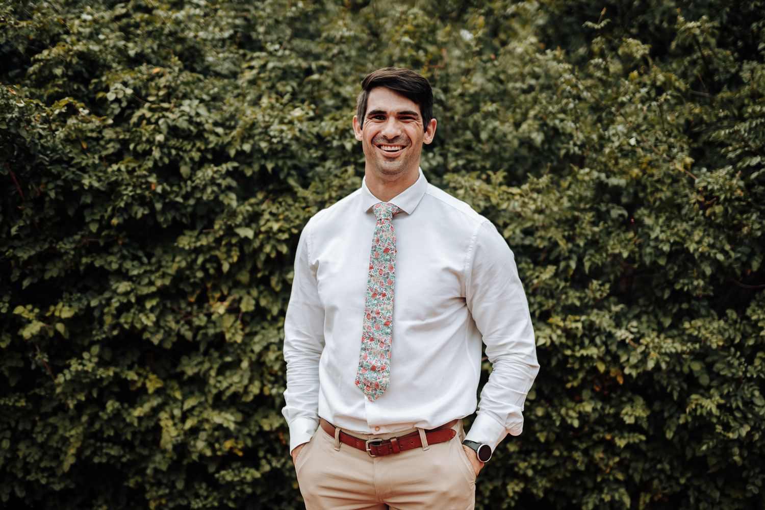 Groomsman in white shirt and floral tie poses confidently against green hedge.
