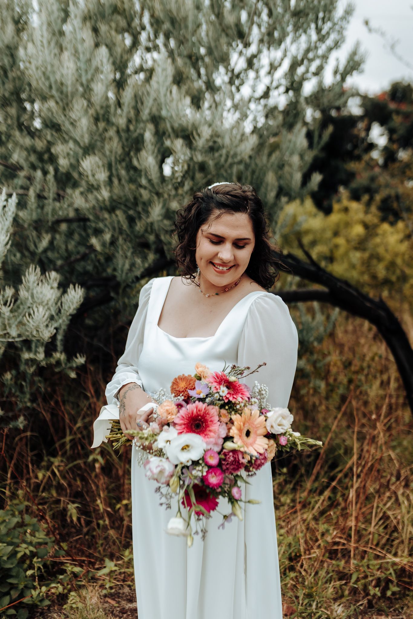 A bouquet of pink and coral flowers complements an elegant white wedding dress in an outdoor setting.