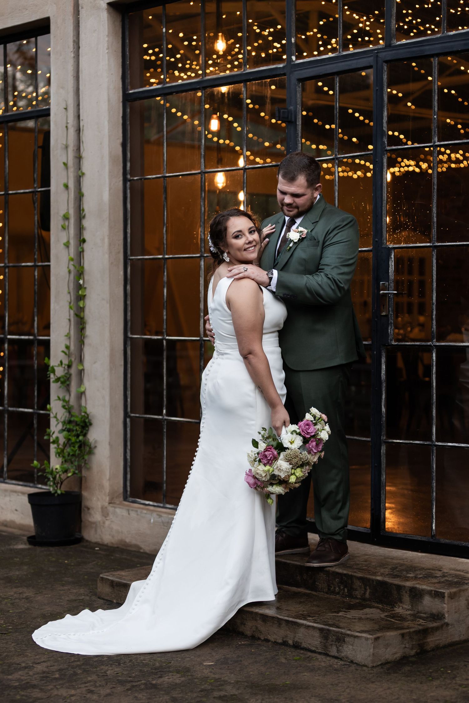 Wedding couple poses outside venue with twinkling lights and industrial windows in background.