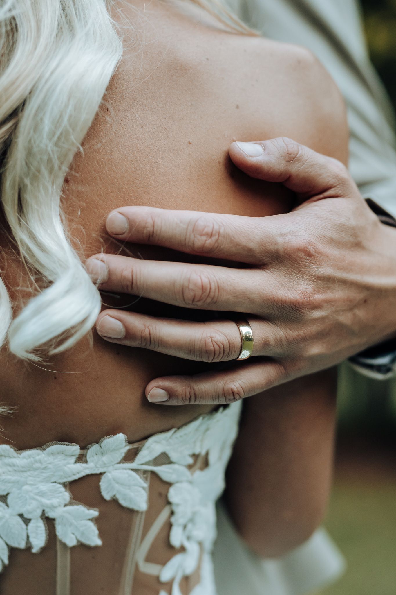 Close-up detail of delicate lace wedding dress with hands wearing gold ring against bare shoulder.