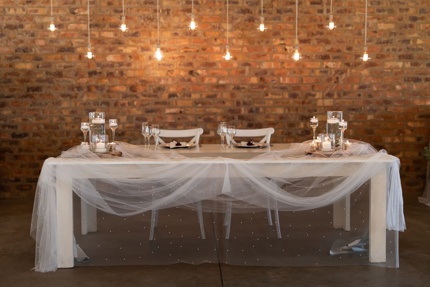 Wedding head table with white tulle draping and hanging lights against exposed brick wall.