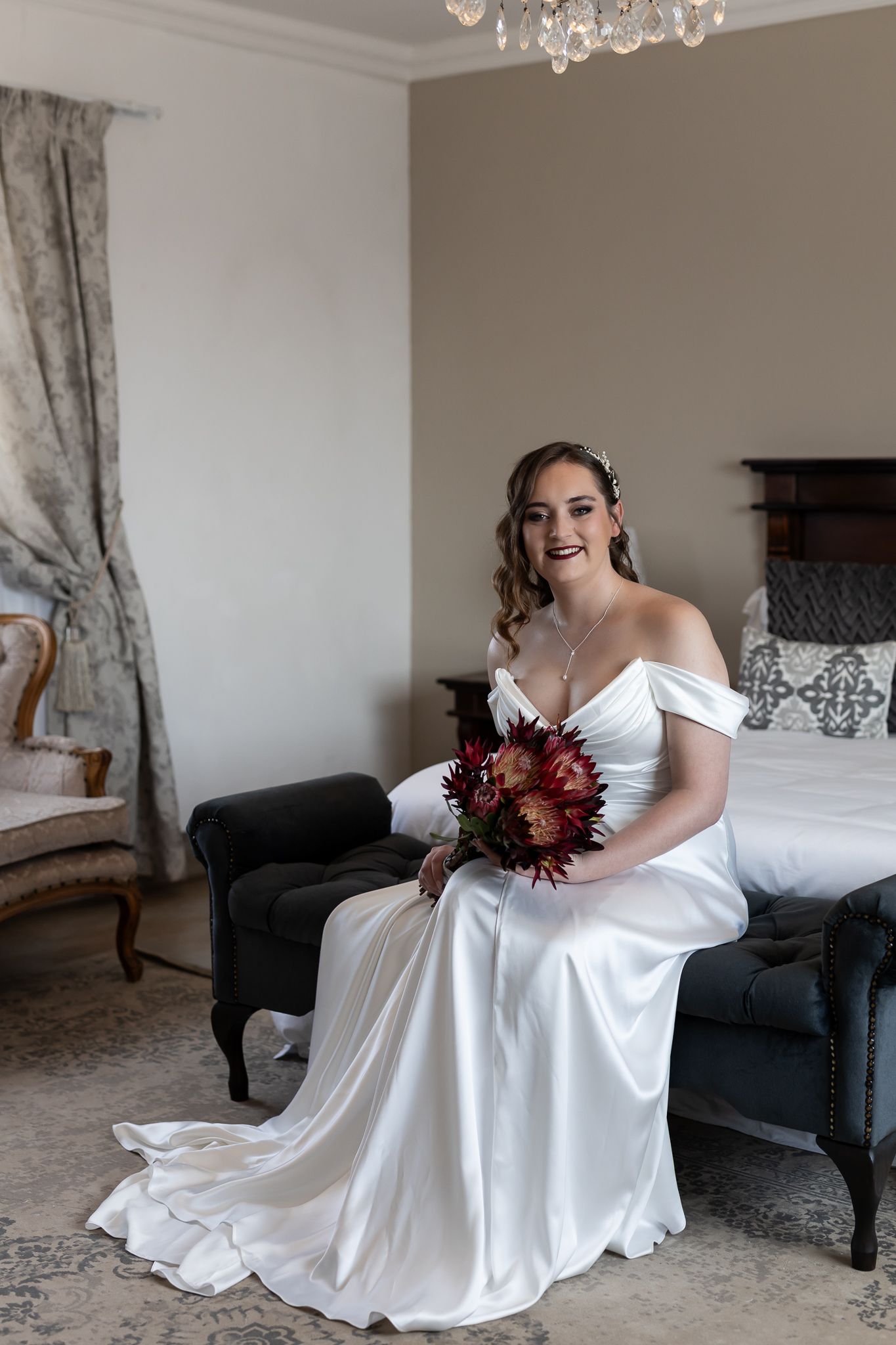 Elegant bride in off-shoulder white gown sitting on bed with dark bouquet in luxurious room.