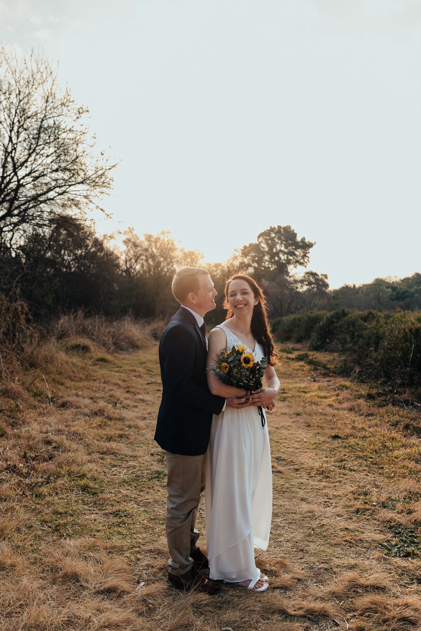 A romantic sunset wedding photo on a hill with warm golden light filtering through the trees.