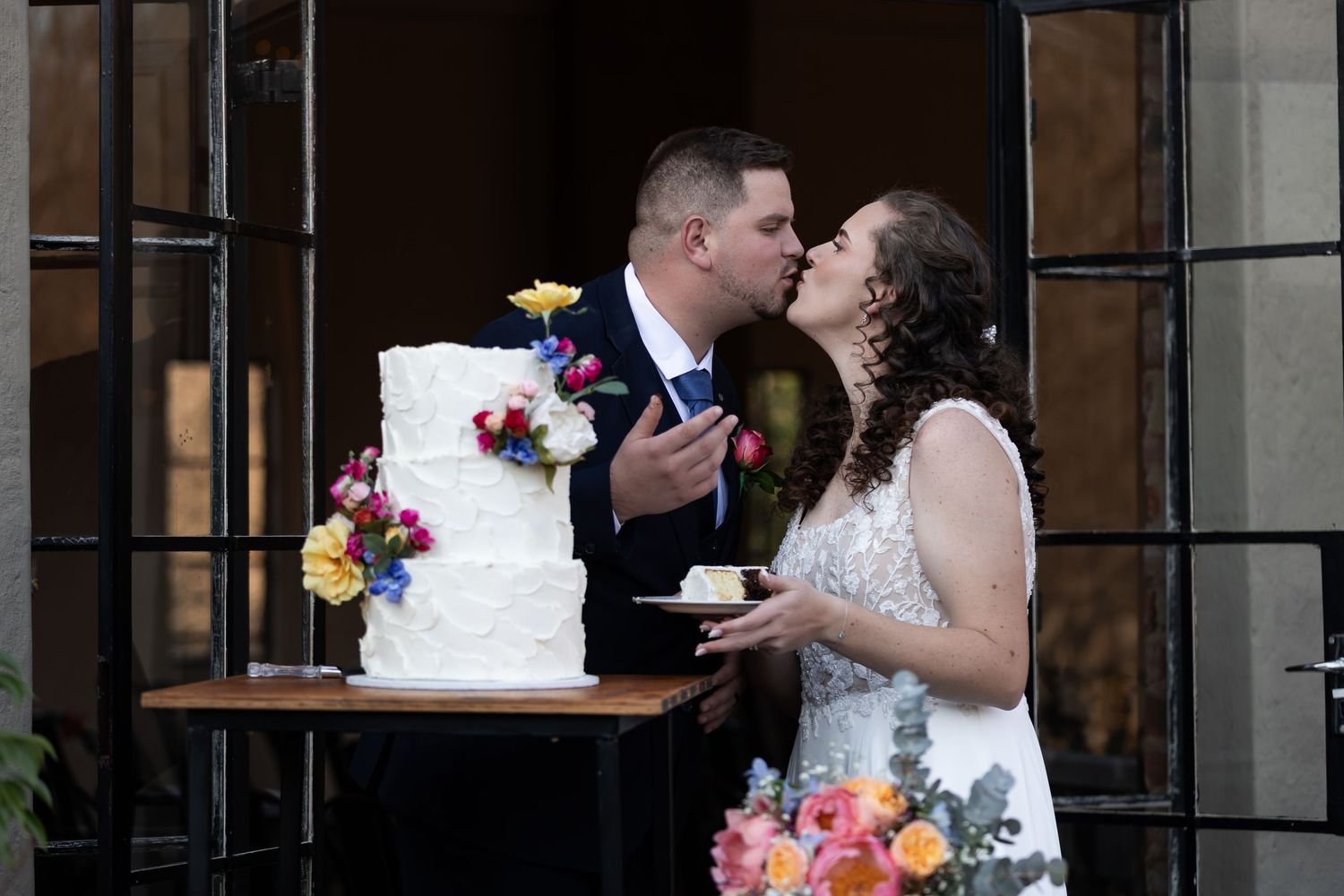 A couple shares a sweet moment while tasting their multi-tiered white wedding cake decorated with colorful flowers.