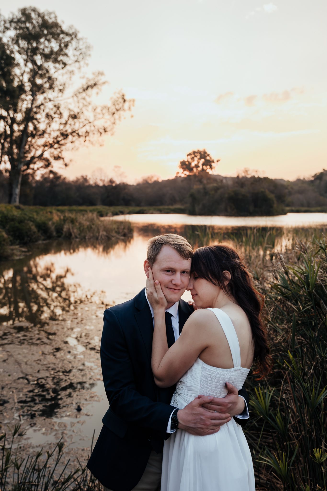 A romantic sunset photoshoot by a serene lake with tall grasses and trees silhouetted against the warm evening sky.