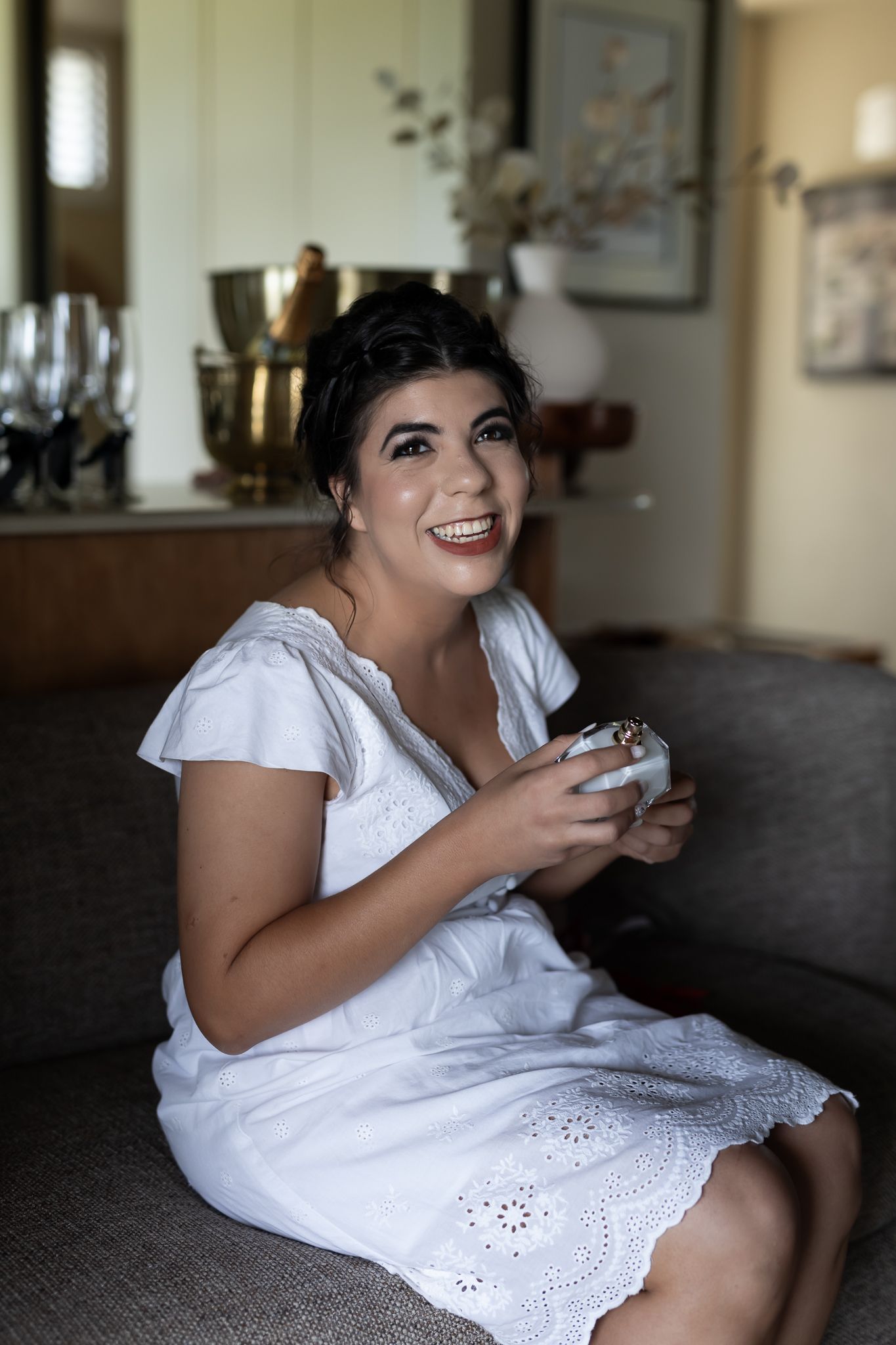 A person in white sits on a dark couch holding something while smiling warmly at the camera.