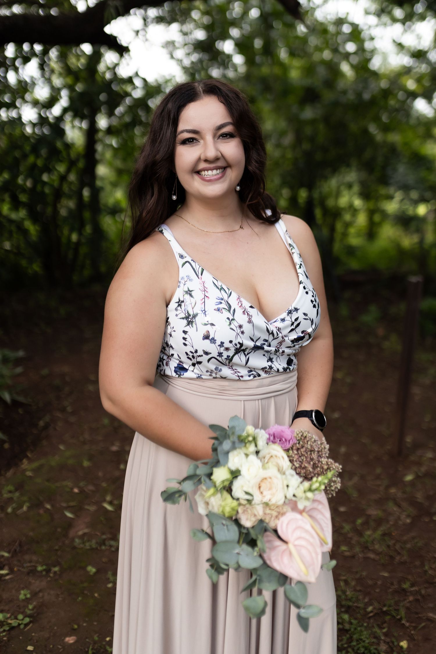 Bridesmaid smiles while holding elegant bouquet in floral dress against green garden background.