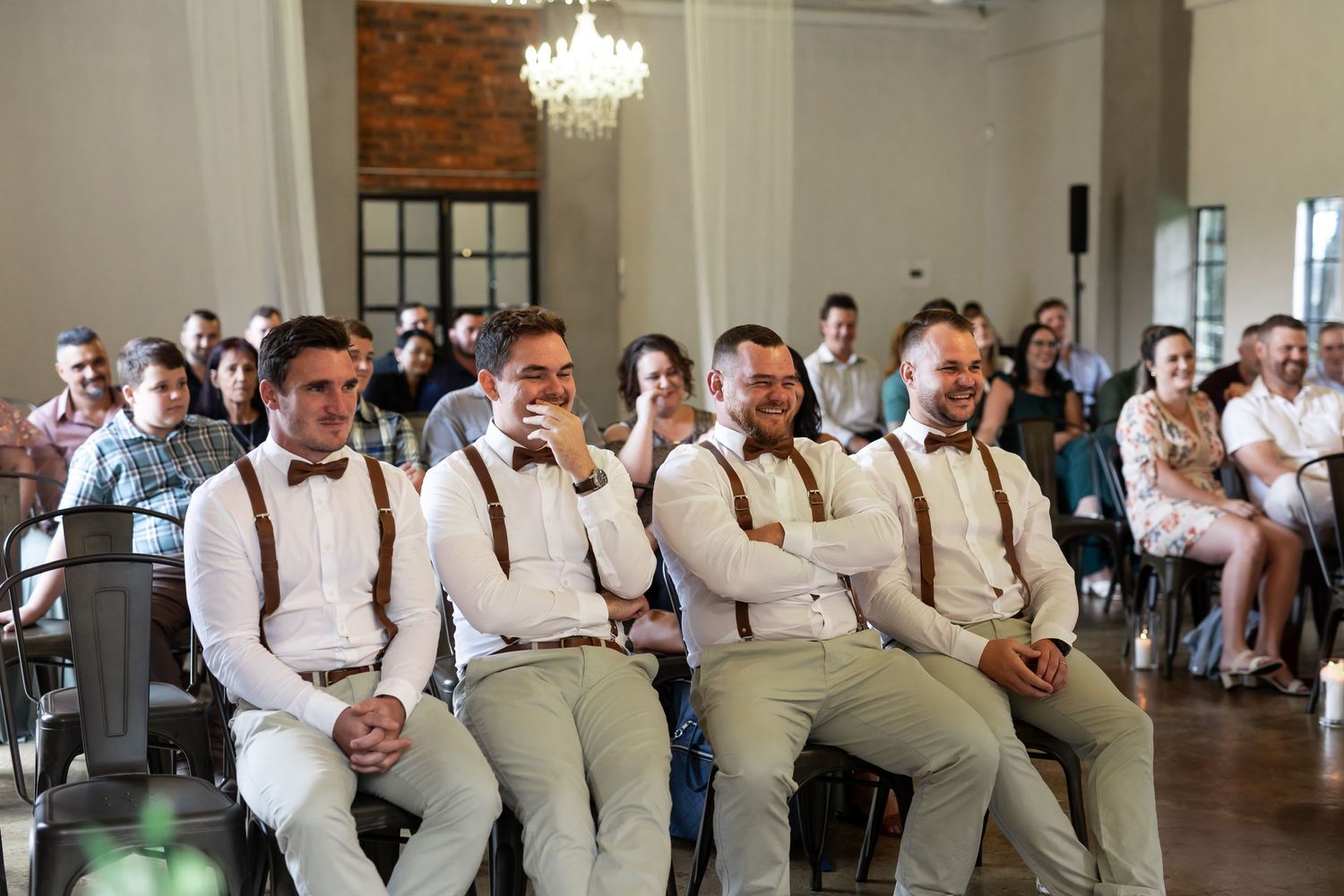 Groomsmen in suspenders and bowties seated in a row during wedding ceremony.