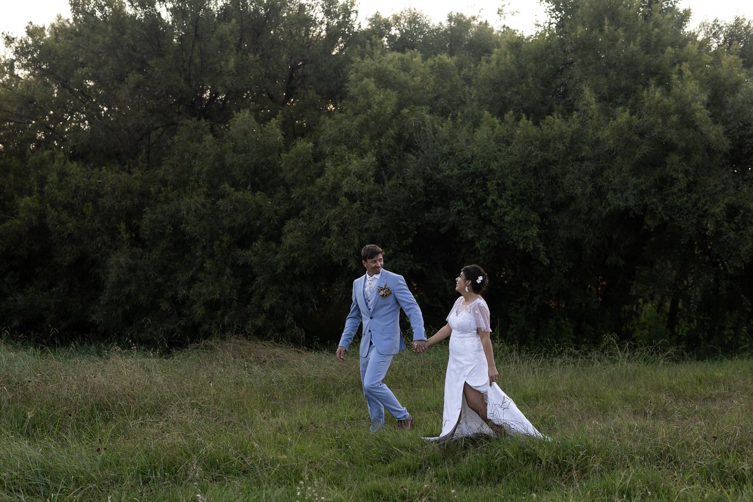 A couple strolls through a green field with trees in the distance.
