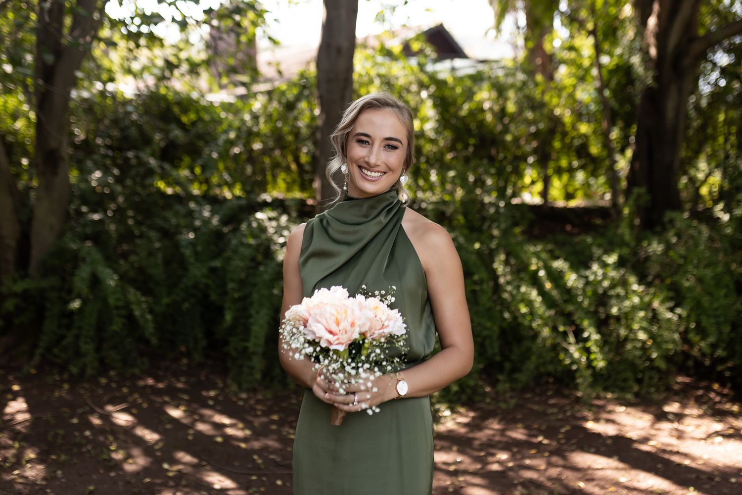 A person in an elegant green dress holds a pink and white bouquet in a sunlit garden.