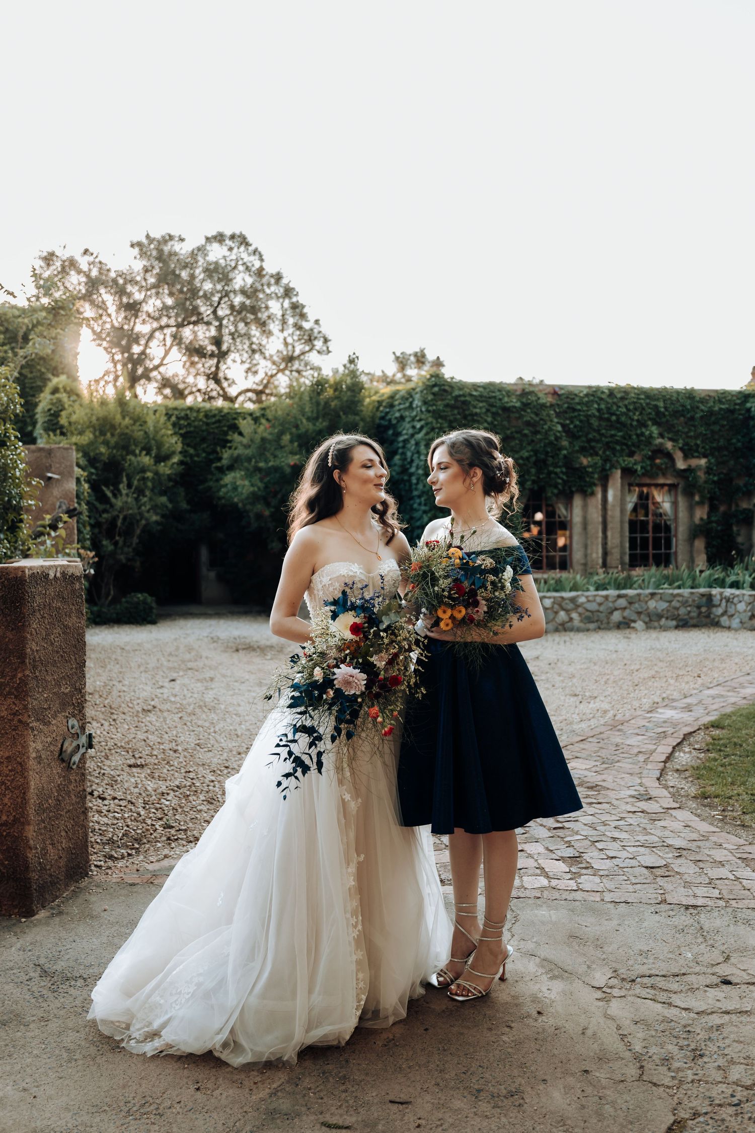 Bride in flowing white gown and bridesmaid in navy share a sunset moment with beautiful bouquets.