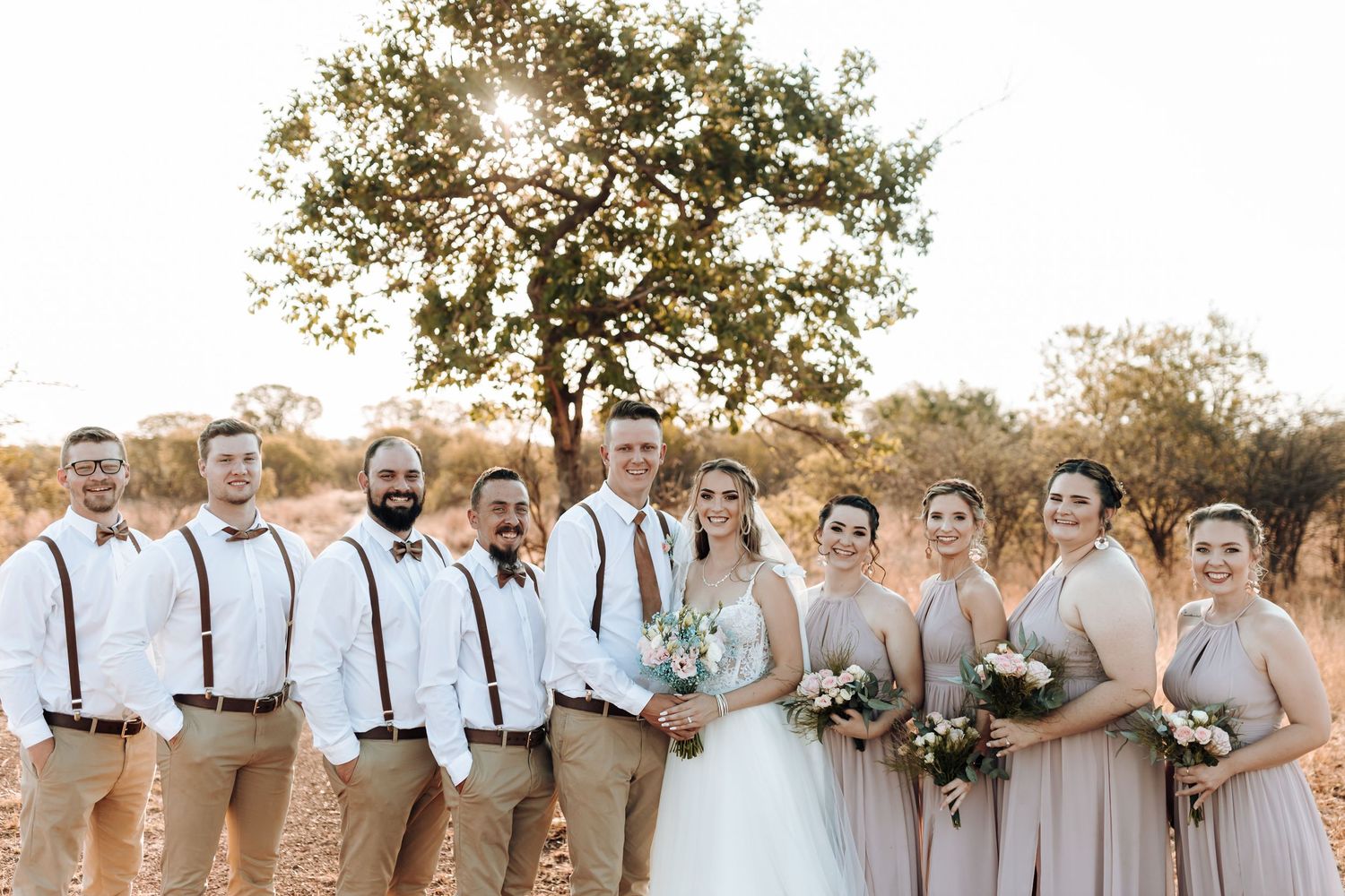 Bridal party in coordinated khaki and blush outfits gathered outdoors for formal photo.