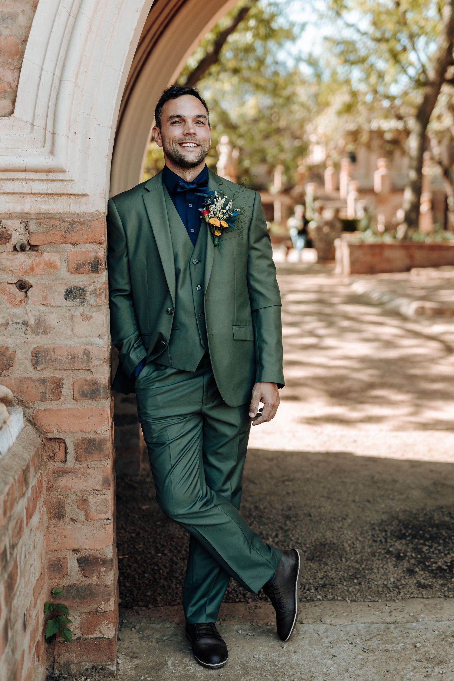 A person in a fitted green suit and boutonniere poses against a brick archway.
