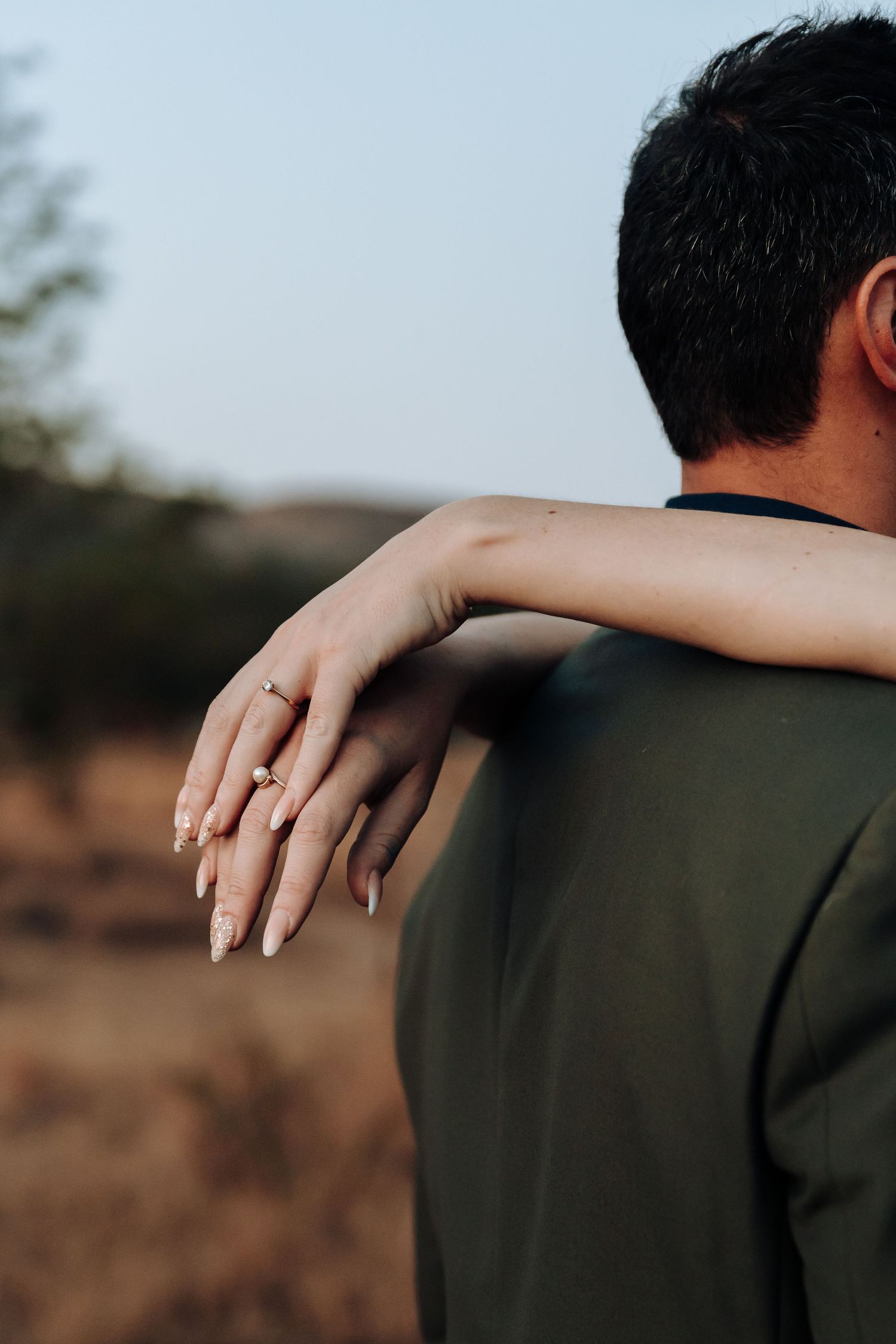 Close-up of bride's hand with engagement ring resting on groom's shoulder against natural backdrop.