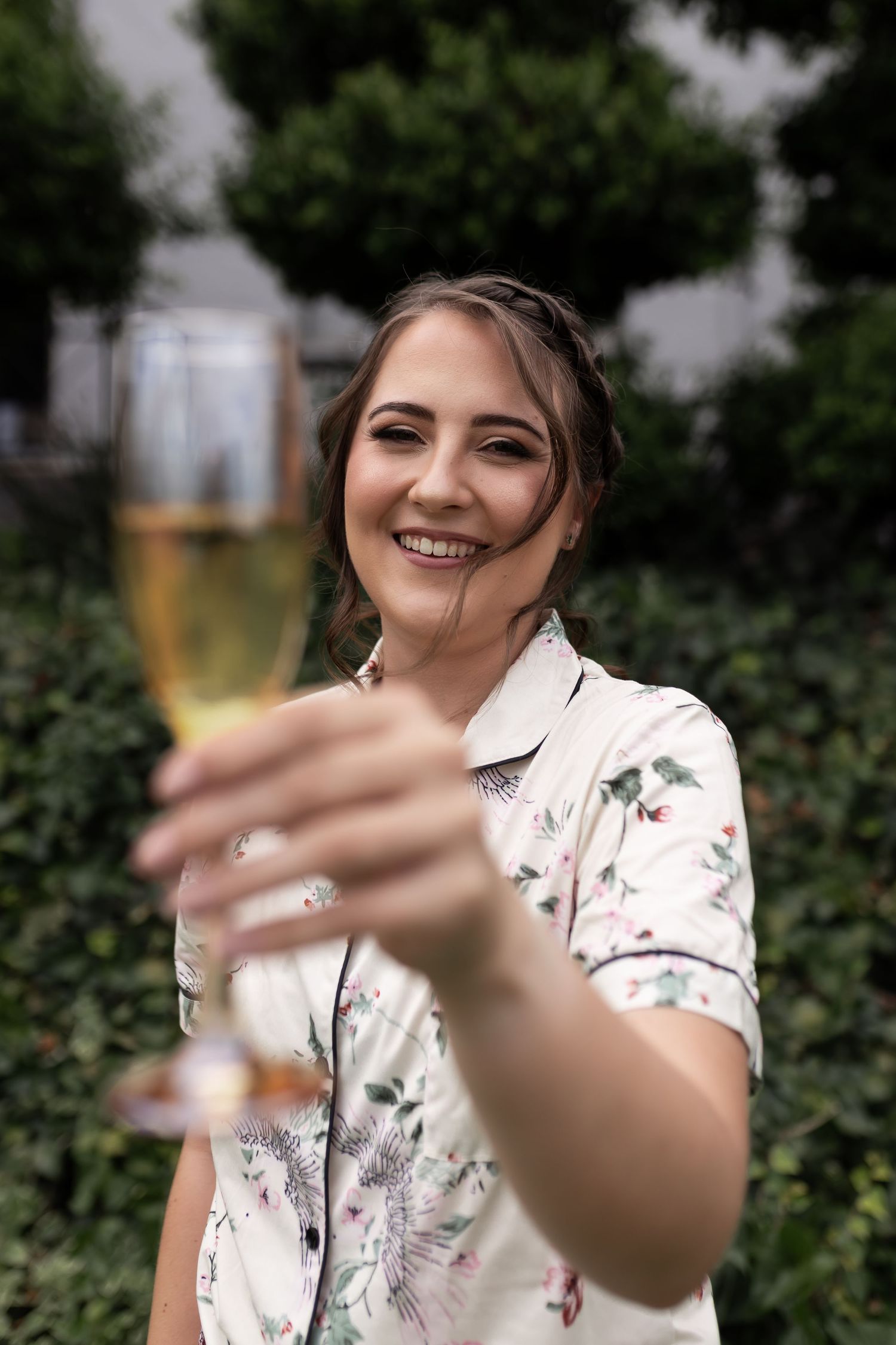 A person in a floral blouse holds up a champagne glass while smiling outdoors.