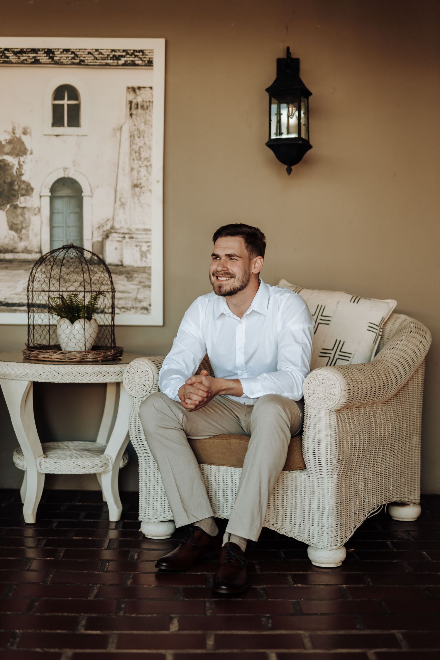 A well-dressed person sits in a wicker armchair next to a decorative side table with vintage lantern decor.