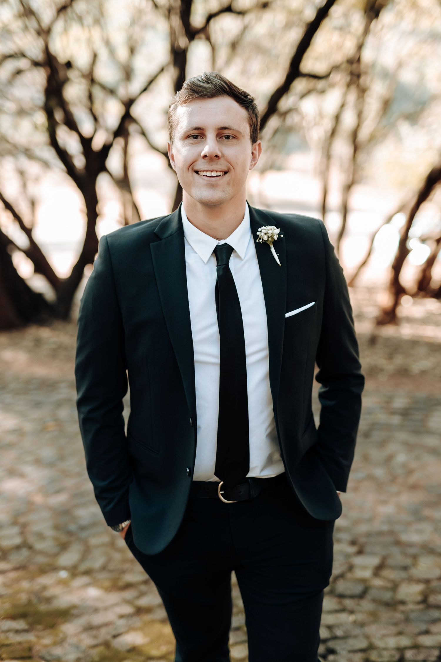 Groomsman in classic black suit smiles while posing on stone path outdoors.