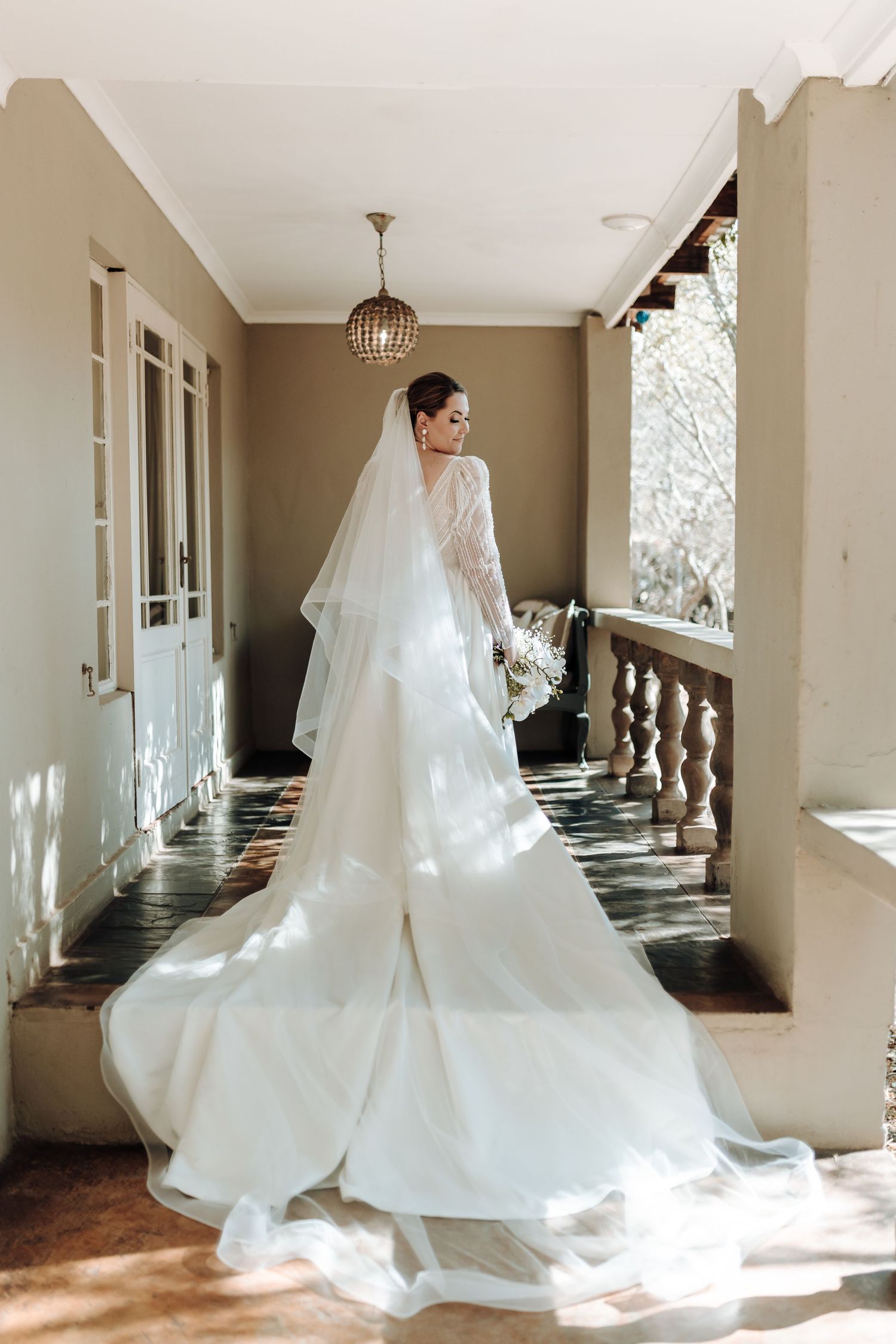 Dramatic backlit bridal portrait showing flowing white wedding dress train on elegant porch.