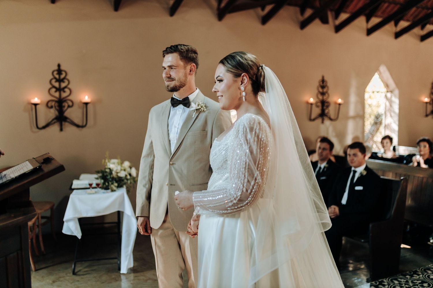 A bride and groom stand together at the altar in a chapel adorned with chandeliers and arched windows.