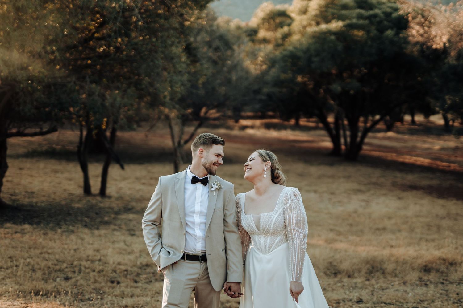 Newlyweds share a romantic moment in a sun-dappled forest setting wearing elegant wedding attire.