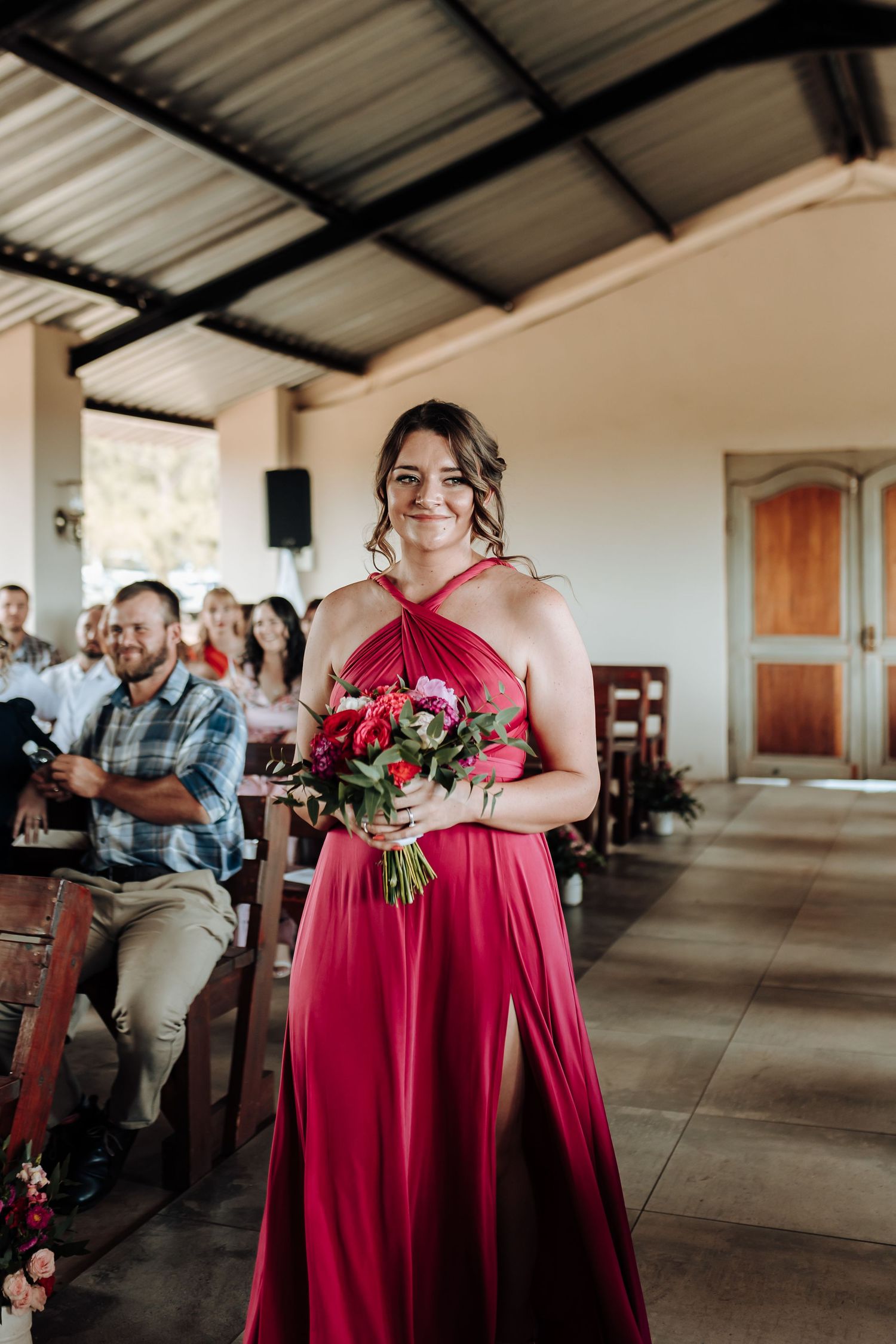 Bridesmaid in flowing red halter dress walks down aisle with pink floral bouquet.
