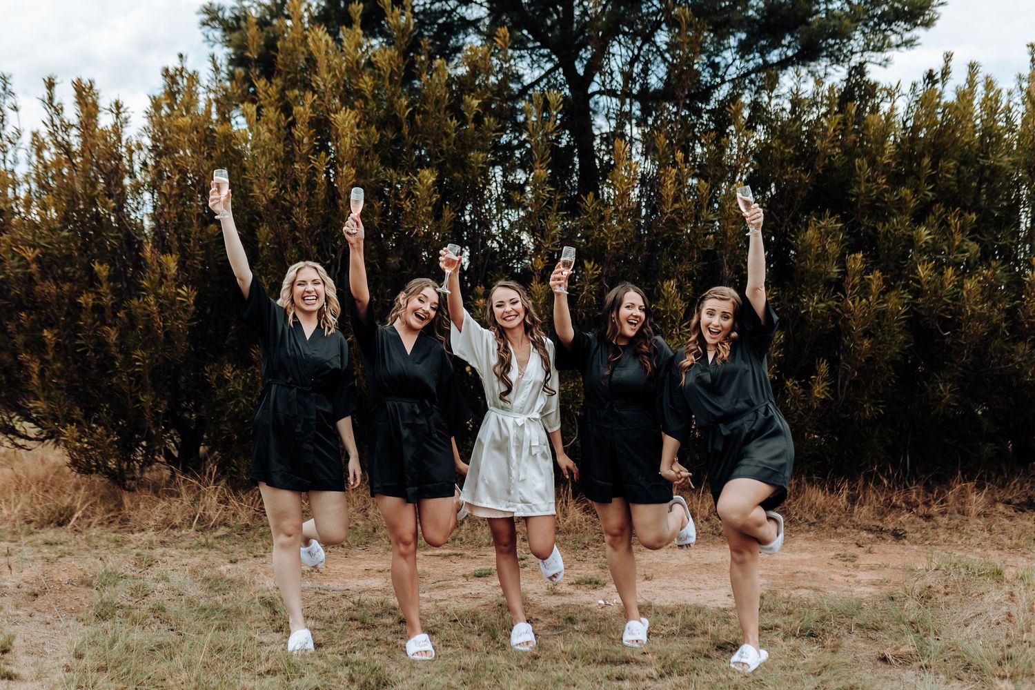 Bridal party in black robes jumps with excitement during an outdoor photo session.