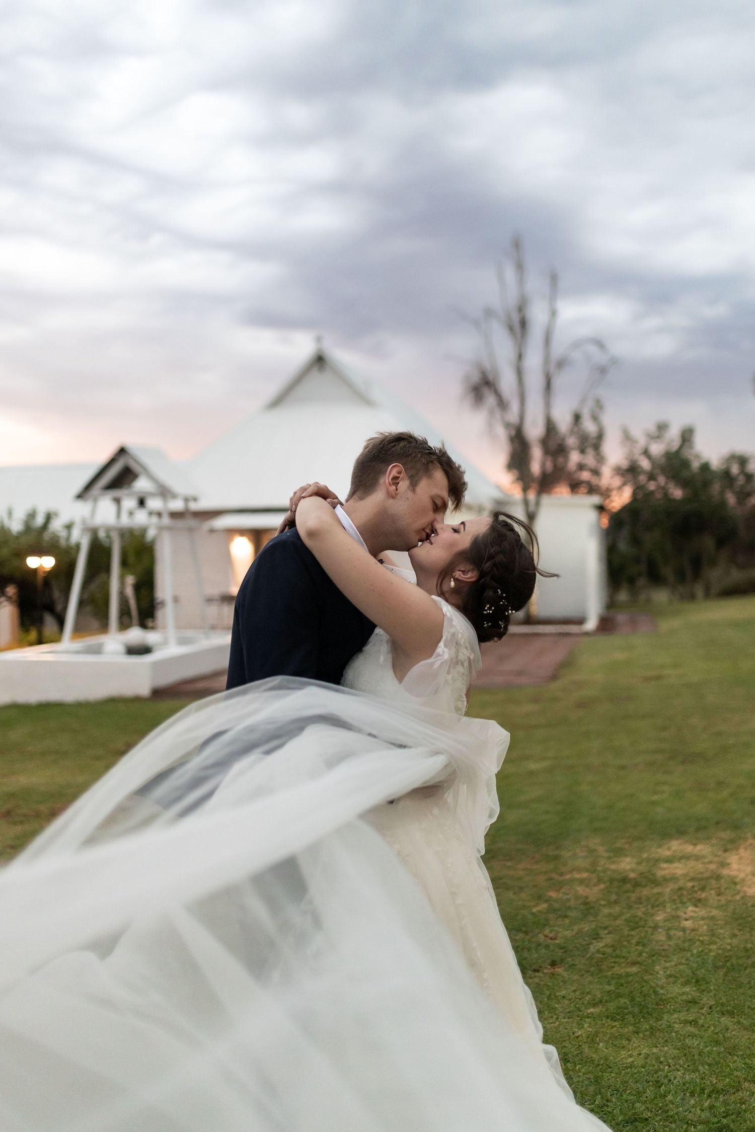A romantic wedding couple dances on a lawn at sunset with a flowing white dress and chapel in the background.