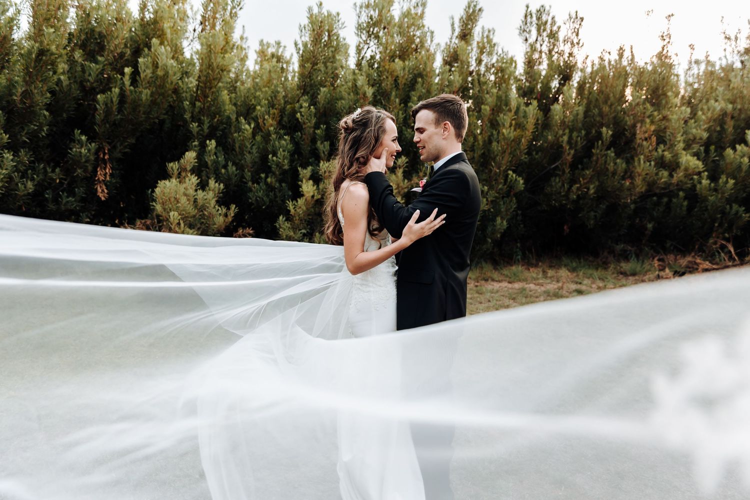 Elegant bride and groom share tender moment as white veil billows in wind against natural backdrop.