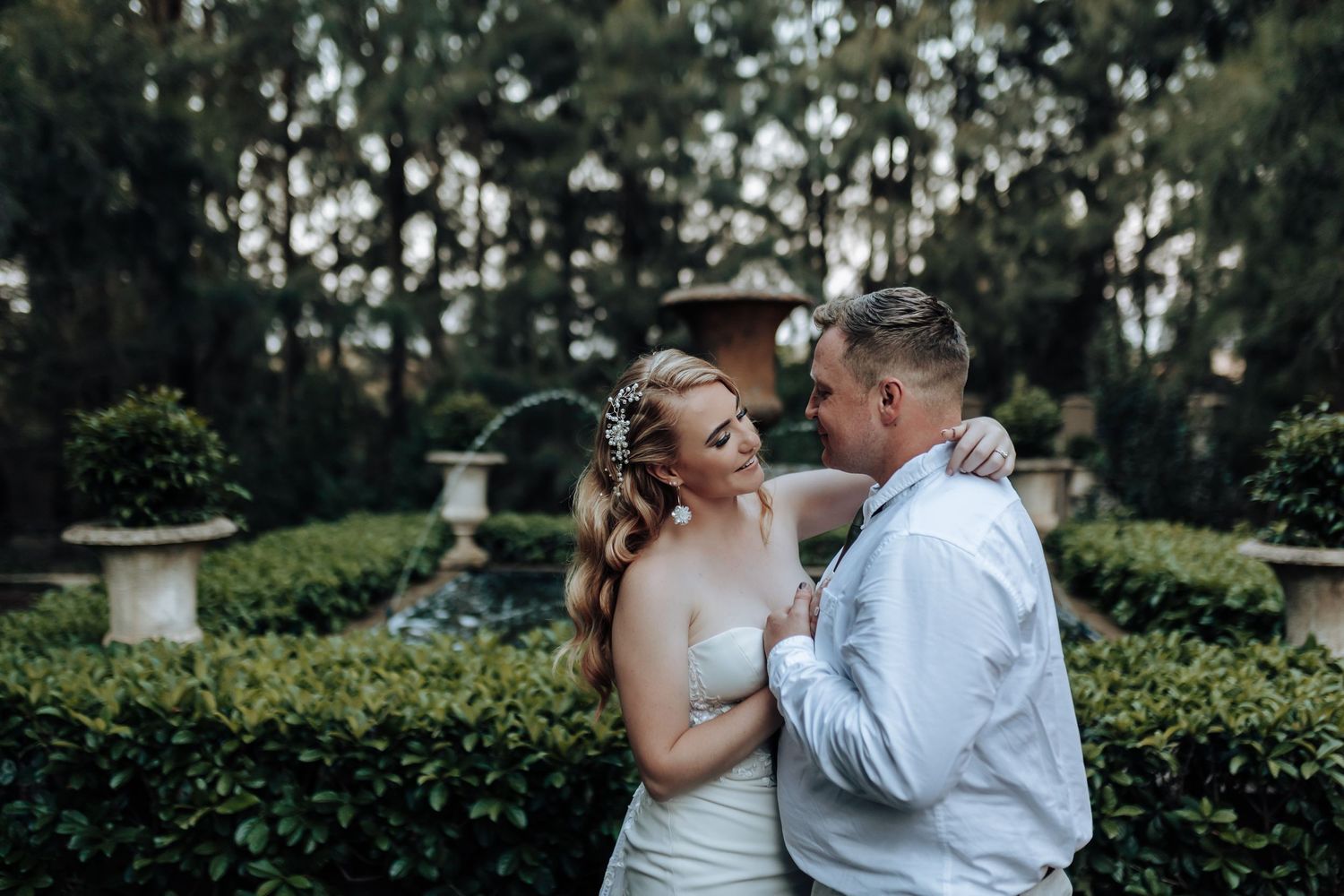 A romantic embrace in a manicured garden with ornamental hedges and pine trees.