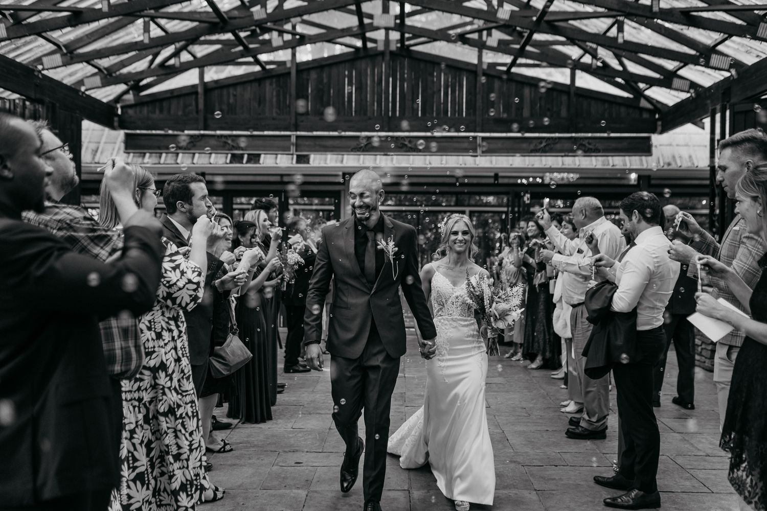 Black and white photo of newlyweds walking through guest-lined aisle at indoor reception.