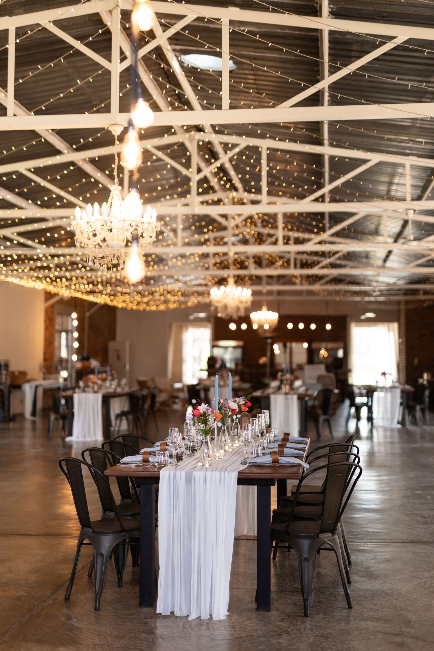 Long rustic reception table with white runner and metal chairs under twinkling string lights in an industrial venue space.