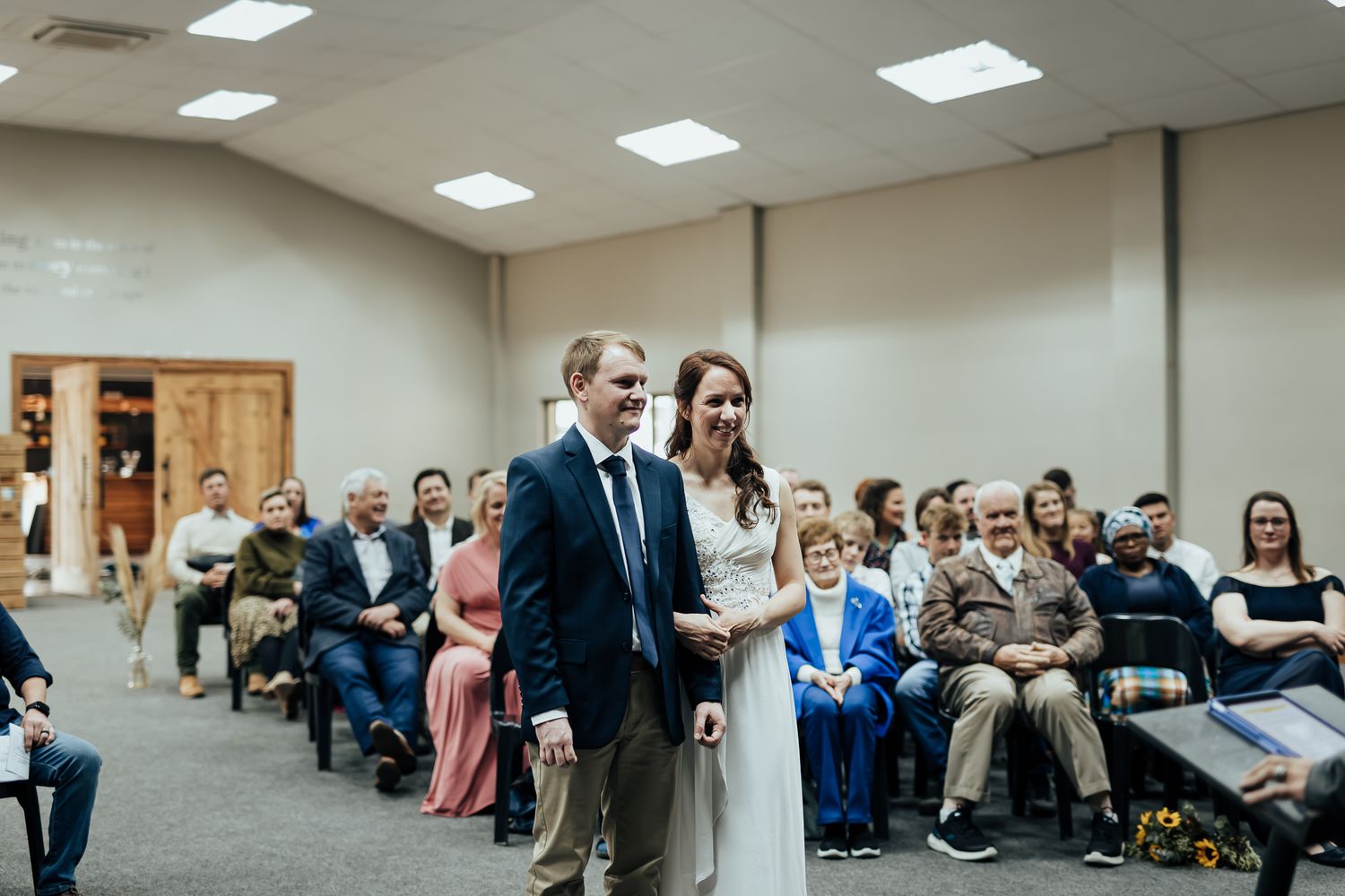 Wide angle view of wedding reception showing guests mingling in a brightly lit community hall.