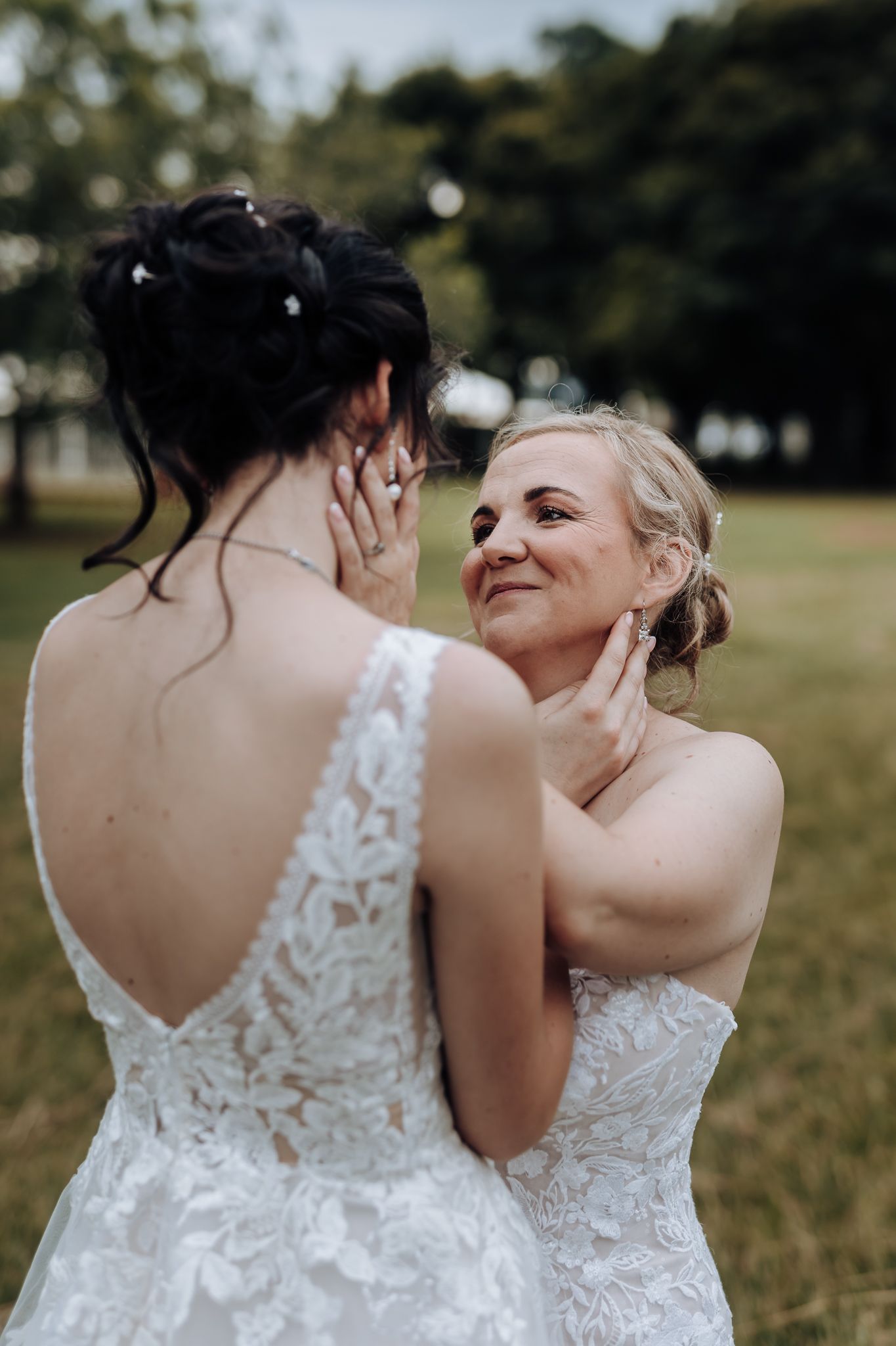 Two brides in lace wedding dresses share a tender moment outdoors.