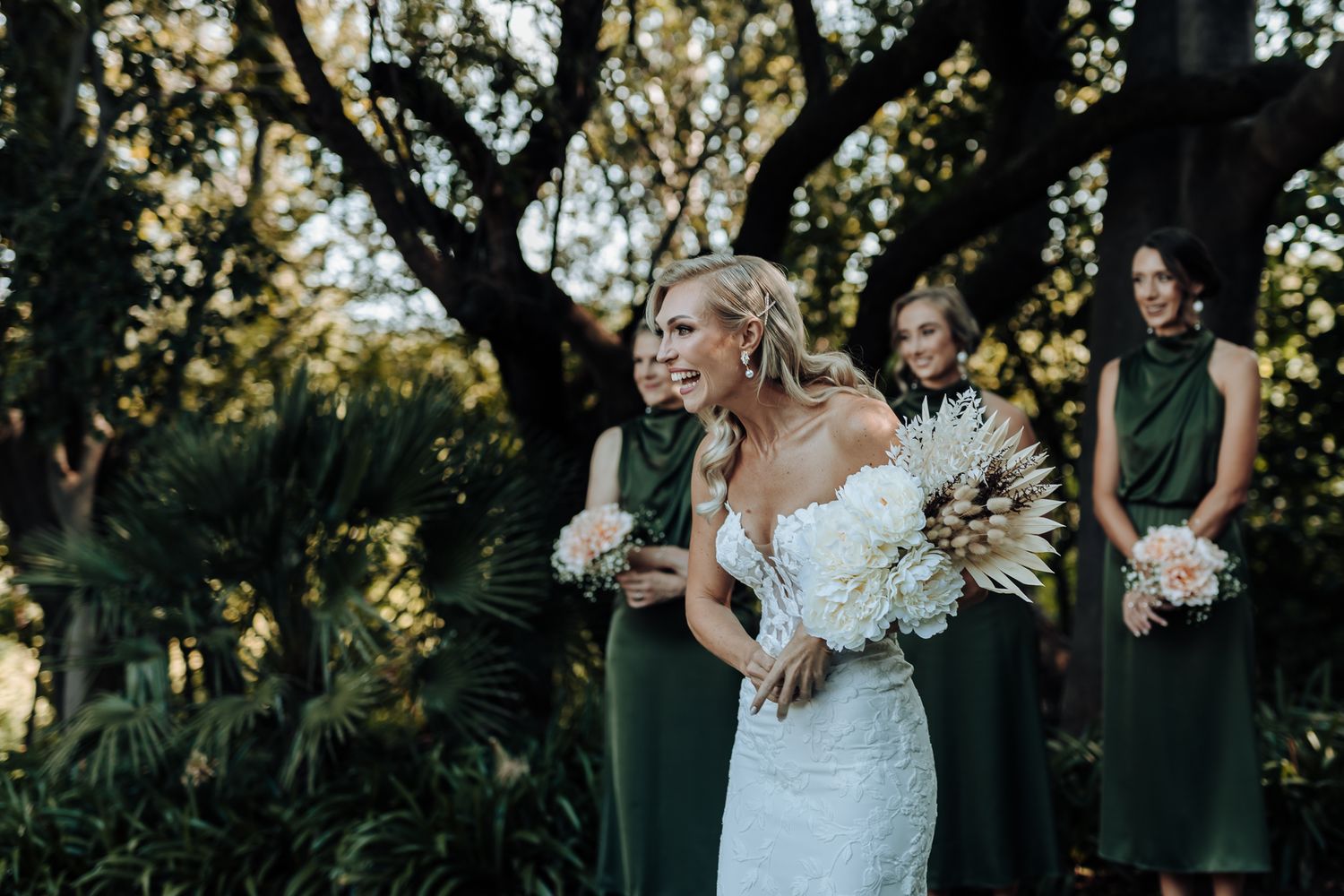 Wedding party in emerald dresses stands behind someone in white carrying a dramatic white and neutral bouquet.