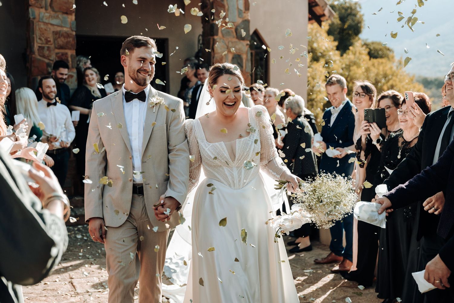 Newlyweds walk through confetti shower outside beautiful stone chapel with mountain backdrop.