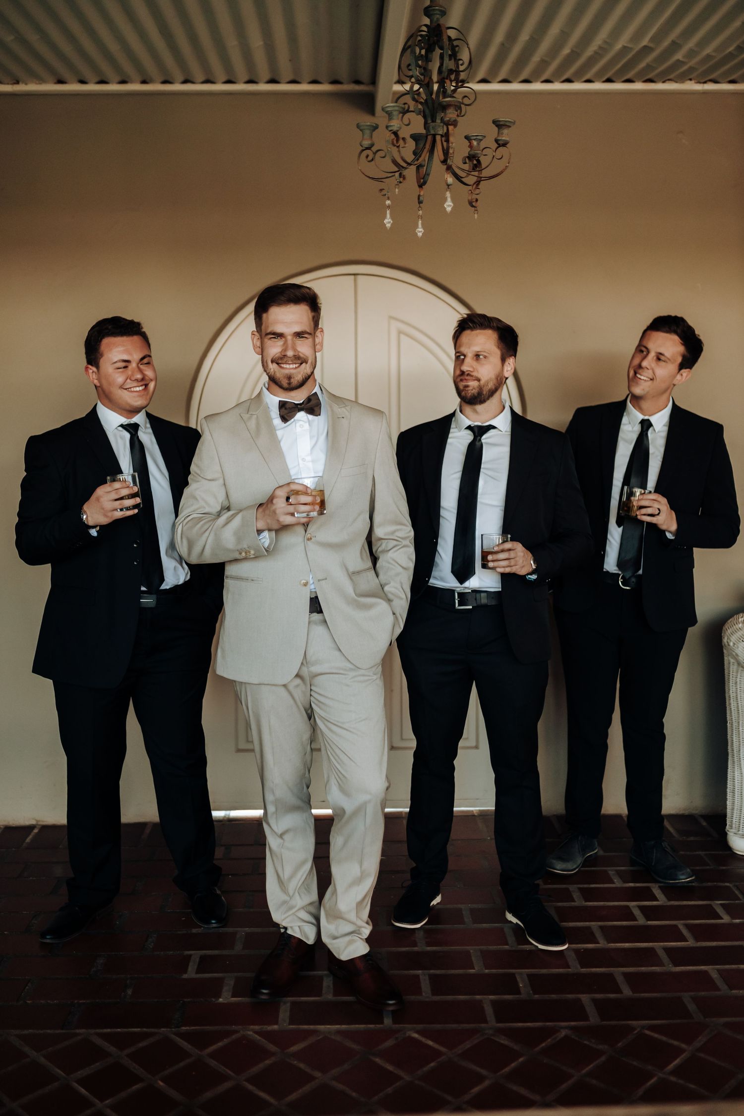 Groomsmen in dark suits flank the groom in beige as they pose with drinks under a chandelier.