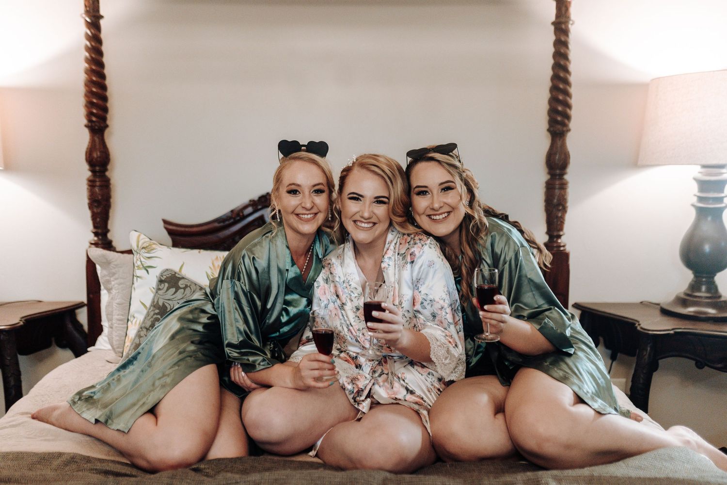 Three friends in matching robes pose together on bed with wine glasses.