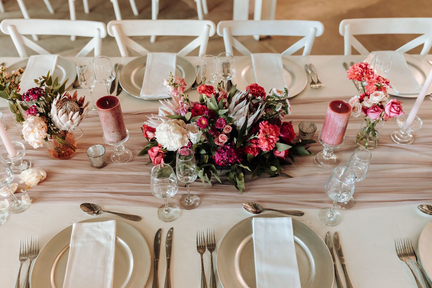 Elegant table setting with pink and red floral centerpieces and white chairs for a wedding reception.