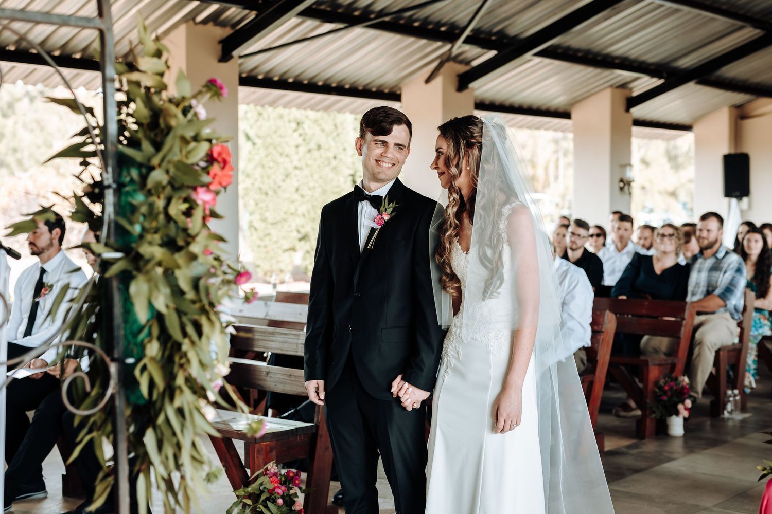 Wedding ceremony takes place under a covered outdoor venue with floral decorations and wooden chairs for guests.